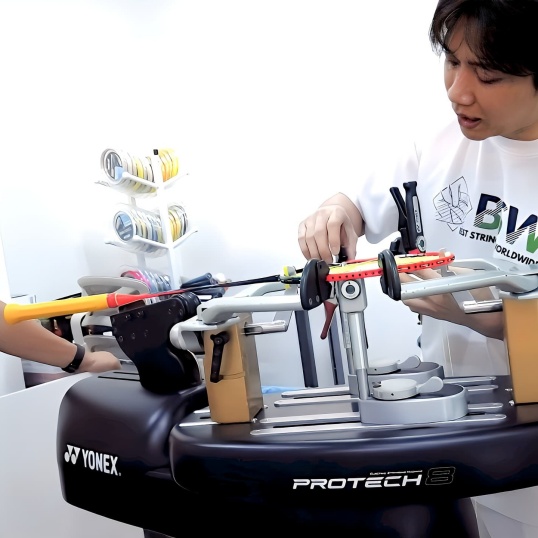 Certified stringer demonstrating precise badminton racket stringing technique on professional Yonex ProTech machine