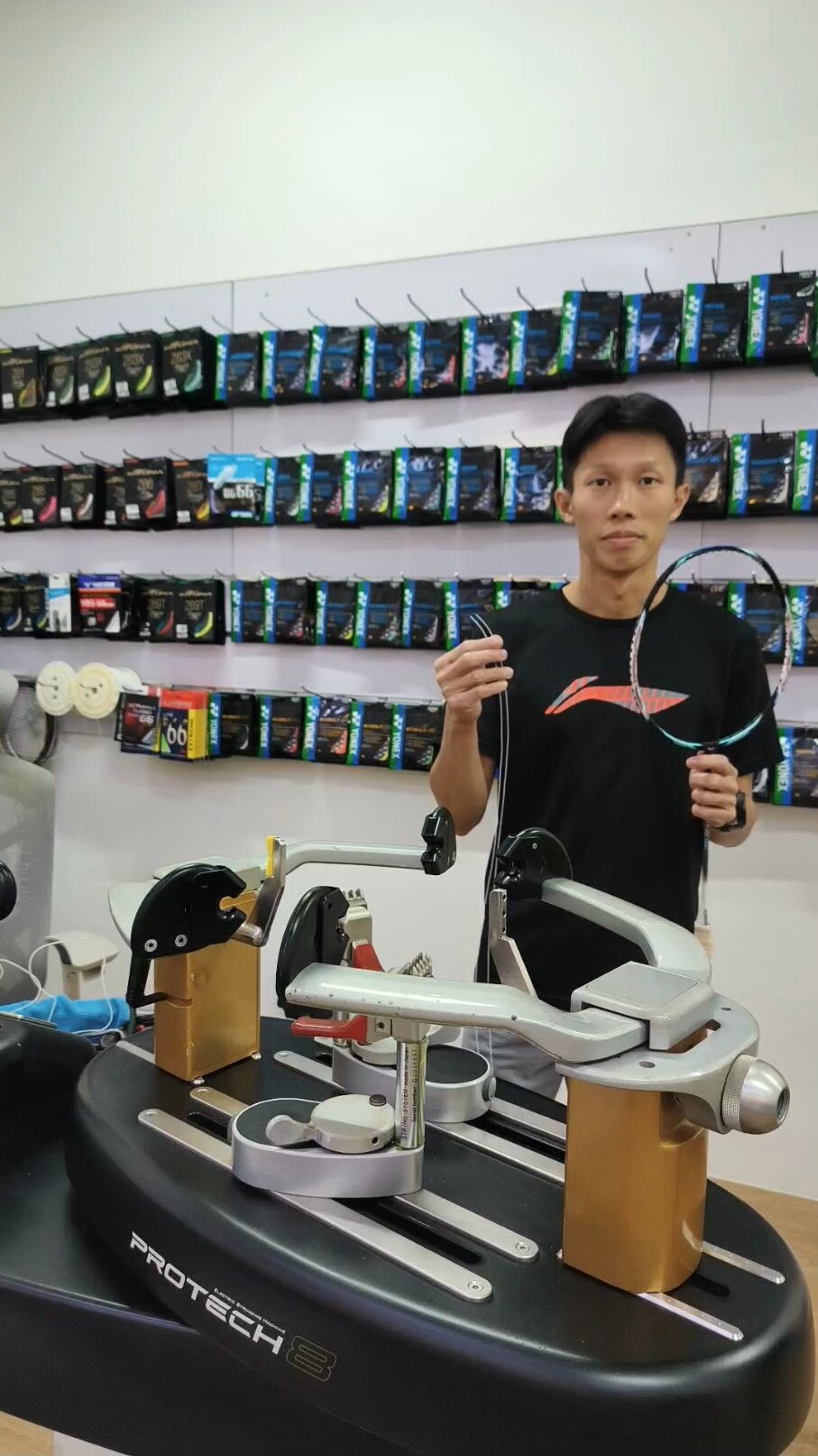 Koh Jian Long Kelvin holding a badminton racquet and loose string beside the stringing machine Koh Jian Long Kelvin holding a badminton racquet and loose string beside the stringing machine
