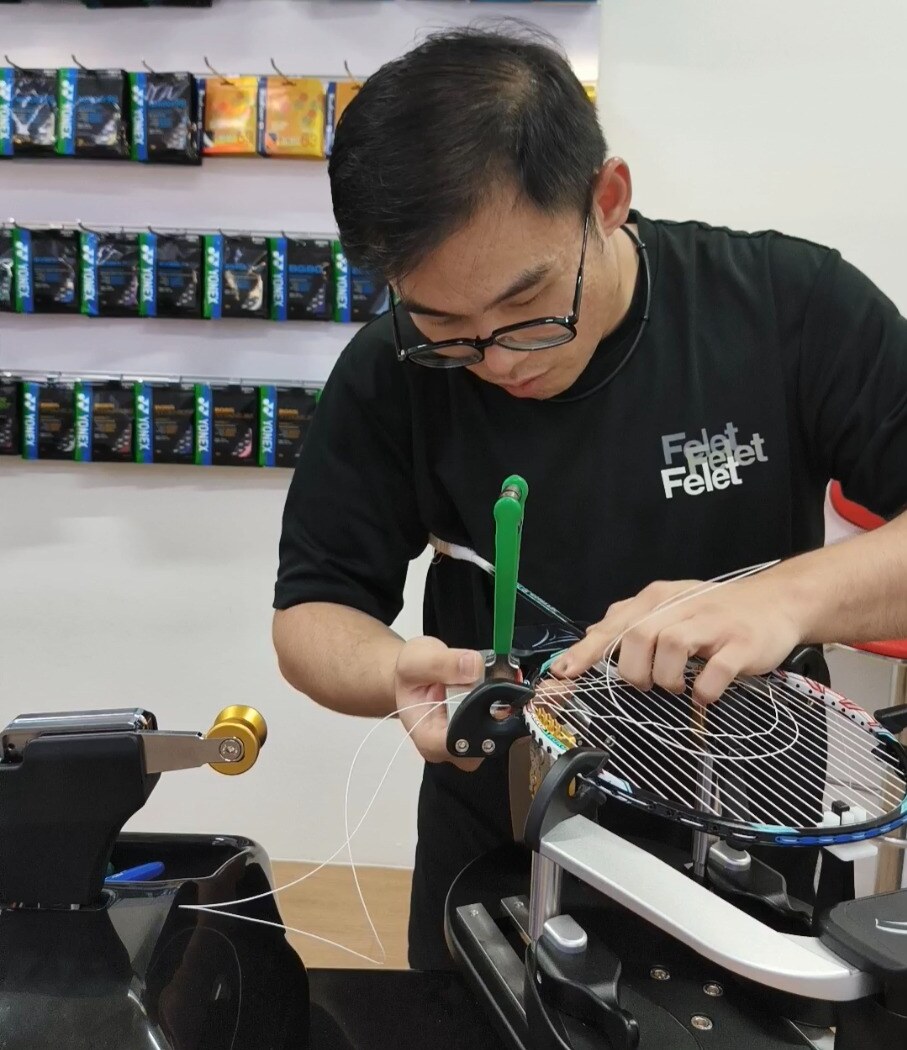 Ng Kim Way guiding string near the badminton racquet frame beside a green-handled clamp Ng Kim Way guiding string near the badminton racquet frame beside a green-handled clamp